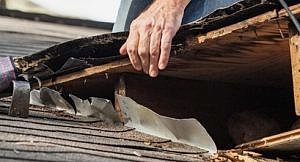 Close up view of man using crowbar and saw to remove rotten wood from leaky roof decking. After removing fascia boards he has discovered that the leak has extended into the beams and decking.